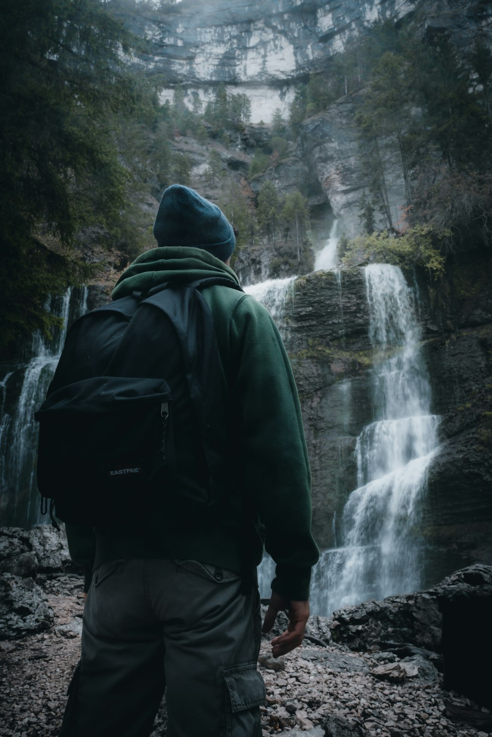 man in green hoodie and black backpack standing in front of waterfalls during daytime