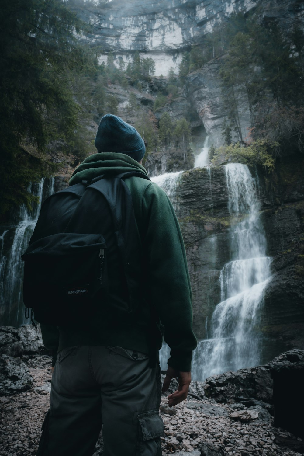 uomo in felpa con cappuccio verde e zaino nero in piedi davanti alle cascate durante il giorno