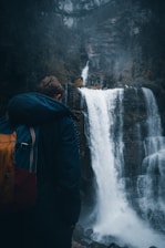 A person wearing a blue jacket and carrying a colorful backpack stands in front of a waterfall. The surrounding area is natural, with dense forest and mist adding a mystical atmosphere.