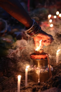 A hand adorned with multiple bangles is carefully lighting a small lamp placed on a brass container. Surrounding the setup are lit candles and incense sticks, with smoke wafting in the air, creating a serene and spiritual ambiance.