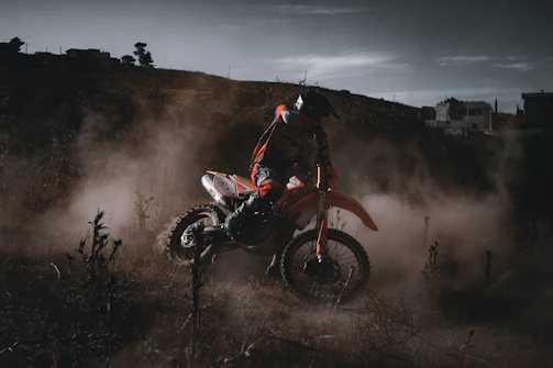 A rugged enduro rider kicking up dust as they navigate a steep mountain trail at sunset.