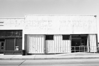 A black and white photograph of an old storefront with a faded sign, showing signs of wear and age. The windows are covered, and the facade has peeling paint. A metal railing and a trash can are visible on the sidewalk in front of the building.