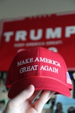A red baseball cap with the phrase 'MAKE AMERICA GREAT AGAIN' embroidered in white letters. An American flag symbol is also present on the side of the cap. A hand is holding the cap, and a red sign with partially visible white lettering is in the background.
