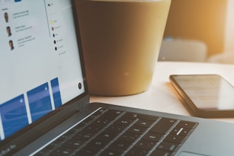 A close-up of a laptop keyboard with a screen showing a user interface, possibly a dashboard. Nearby, a smartphone lies on a light-colored surface, partially in shadow. A large plant pot is visible in the background, bathed in warm sunlight.