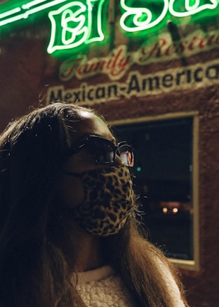 A person wearing a leopard print face mask and sunglasses stands in front of a neon-lit sign that reads 'El Sol' and 'Mexican-American.' The image has a nighttime setting with a dimly lit ambiance.