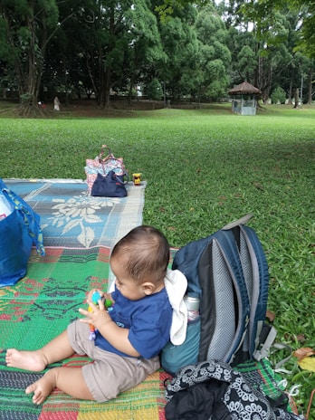A baby is sitting on a colorful mat in a park, playing with a toy. Surrounding the baby, there are bags and a backpack on the mat. The park features lush green grass and tall, dense trees in the background, creating a peaceful natural setting.