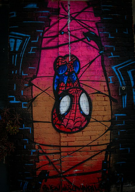 A vibrant graffiti art piece on a brick wall depicting a cartoon-like character resembling a superhero in a red and blue suit, hanging upside down with a web. The background features a mix of pink, black, and orange tones with abstract lines and building outlines.