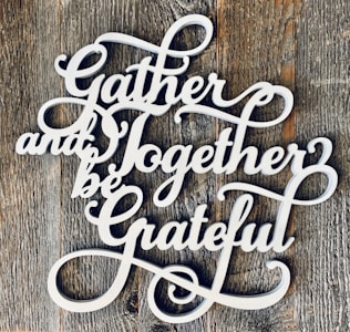 Decorative white cursive text spells out 'Gather and Together be Grateful' against a rustic wooden background.