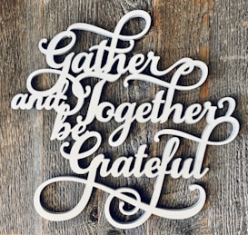 Decorative white cursive text spells out 'Gather and Together be Grateful' against a rustic wooden background.