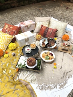 An outdoor picnic setup features a variety of intricately decorated pillow cushions in earthy tones and warm colors, placed on textured blankets and throws. There are a number of white decorative boxes and vibrant marigold flower arrangements scattered around. The setup includes several plates and trays with an assortment of desserts, crackers, and dips. The ground is made of stone tiles, adding a rustic feel to the overall scene.