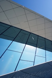 A modern building facade featuring large reflective glass panels and geometric patterns. The upper part includes an overhanging roof structure with a tiled design. The sky is clear, contributing to the structure's clean and sharp appearance.