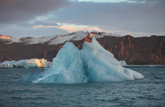 An iceberg floating in the ocean with snow-capped mountains in the background. The sky is partially cloudy with the sun casting a warm light on the landscape.