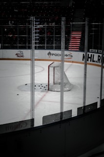 The image captures an empty ice hockey rink, focusing on a single goal with multiple hockey pucks scattered in front of it. The rink is surrounded by transparent protective boards with advertisements visible in the background.