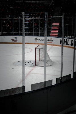 The image captures an empty ice hockey rink, focusing on a single goal with multiple hockey pucks scattered in front of it. The rink is surrounded by transparent protective boards with advertisements visible in the background.
