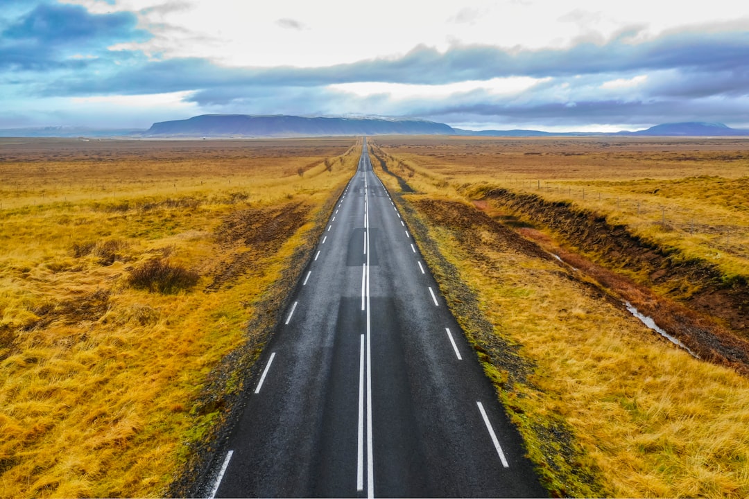 black asphalt road between brown grass field under blue and white sunny cloudy sky during daytime