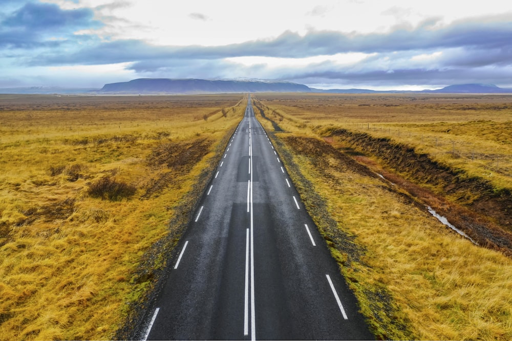 black asphalt road between brown grass field under blue and white sunny cloudy sky during daytime