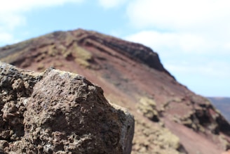 Rugged volcanic rocks are shown in the foreground with a large, sloping terrain that resembles a volcanic landscape in the background. The sky is partly cloudy, and the colors of the terrain range from dark browns and reds to lighter beige tones.