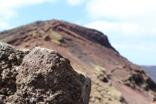 Rugged volcanic rocks are shown in the foreground with a large, sloping terrain that resembles a volcanic landscape in the background. The sky is partly cloudy, and the colors of the terrain range from dark browns and reds to lighter beige tones.