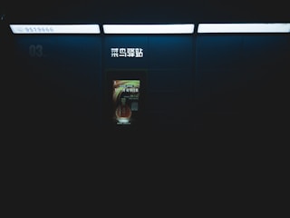 A dimly lit setting featuring a wall with an illuminated advertisement and some signage in Chinese characters. The surrounding area is mostly dark, suggesting an indoor location like a subway or train station. The advertisement has a bottle image with text.