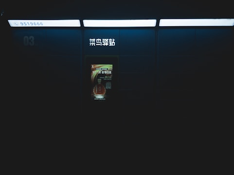 A dimly lit setting featuring a wall with an illuminated advertisement and some signage in Chinese characters. The surrounding area is mostly dark, suggesting an indoor location like a subway or train station. The advertisement has a bottle image with text.