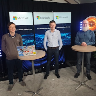 Three people are standing in front of a backdrop with Microsoft branding and the text 'Cloud stories from Norway.' Each person is positioned behind a tall round table. One table has a laptop covered in various tech-related stickers. The setting appears to be a professional or tech-related event with lights on either side.