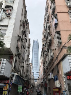 Tall modern skyscraper visible between two older, densely built apartment blocks with multiple air conditioning units. The street below is narrow and bustling, featuring various small shops with signs in a mix of languages. The contrast between the sleek skyscraper and the aged residential buildings is stark.