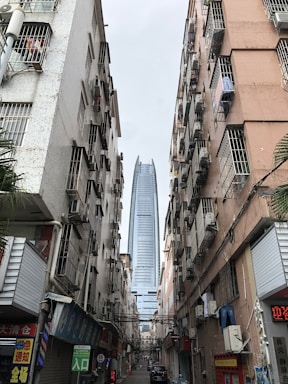 Tall modern skyscraper visible between two older, densely built apartment blocks with multiple air conditioning units. The street below is narrow and bustling, featuring various small shops with signs in a mix of languages. The contrast between the sleek skyscraper and the aged residential buildings is stark.