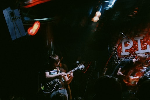 A live musical performance takes place in a dimly lit venue. A guitarist is playing in the foreground with intense focus. In the background, a drummer is energetically engaged with a drum set. A sign with large letters partially reading 'PL' is visible. The lighting is moody with red and blue highlights, creating a dynamic atmosphere.