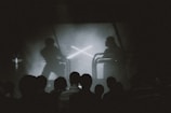 A group of people is silhouetted against a screen showing a dramatic scene. The screen depicts two figures engaged in a sword fight, reminiscent of a popular science fiction saga. The lighting is dim and monochrome, enhancing the focus on the screen's action.