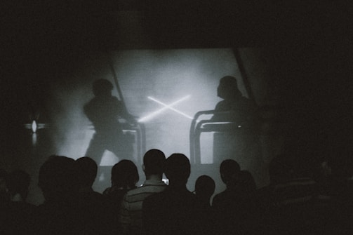 A group of people is silhouetted against a screen showing a dramatic scene. The screen depicts two figures engaged in a sword fight, reminiscent of a popular science fiction saga. The lighting is dim and monochrome, enhancing the focus on the screen's action.