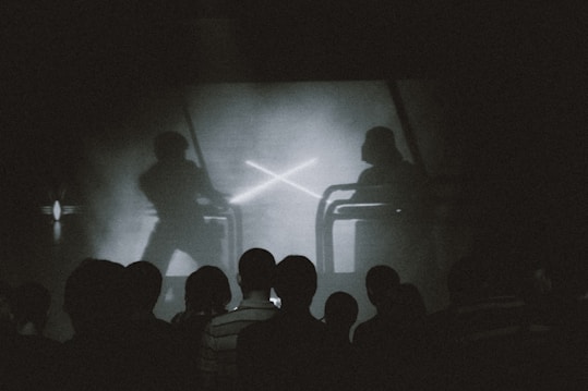 A group of people is silhouetted against a screen showing a dramatic scene. The screen depicts two figures engaged in a sword fight, reminiscent of a popular science fiction saga. The lighting is dim and monochrome, enhancing the focus on the screen's action.