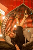 A person is lounging comfortably in a hanging chair with their legs playfully lifted. The setting is warmly lit with a glowing ambiance, and the individual is dressed in stylish attire. The background features a red wall with an interesting pattern that complements the cozy environment.