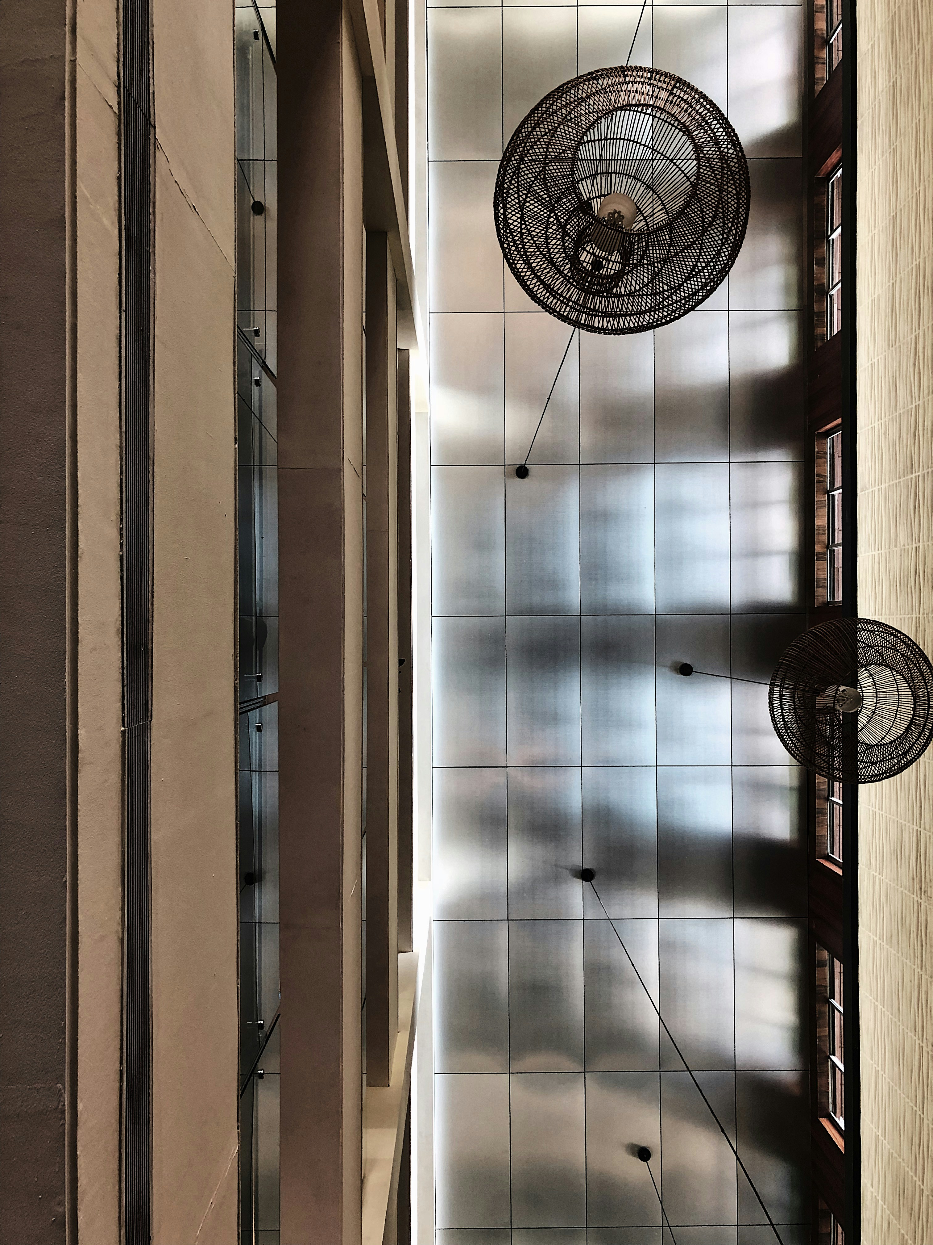 A modern architectural interior view focusing on a high ceiling with visible grid-like panels. Large, decorative wire mesh structures hang from the ceiling, casting intricate shadows. The tall, narrow windows on the sides allow natural light to illuminate the space, creating a geometric and symmetric pattern.