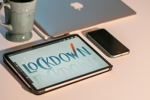 A tablet displaying the word 'LOCKDOWN' with an upward arrow, sitting on a desk alongside a smartphone, a closed laptop featuring a bitten apple logo, and a ceramic mug. The setting appears to be a workspace or home office.