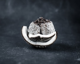 A sliced coconut shell filled with a stack of coconut-covered chocolate balls with chocolate shavings on top, set against a dark background.