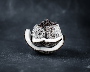 A sliced coconut shell filled with a stack of coconut-covered chocolate balls with chocolate shavings on top, set against a dark background.