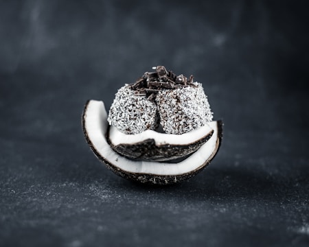 A sliced coconut shell filled with a stack of coconut-covered chocolate balls with chocolate shavings on top, set against a dark background.
