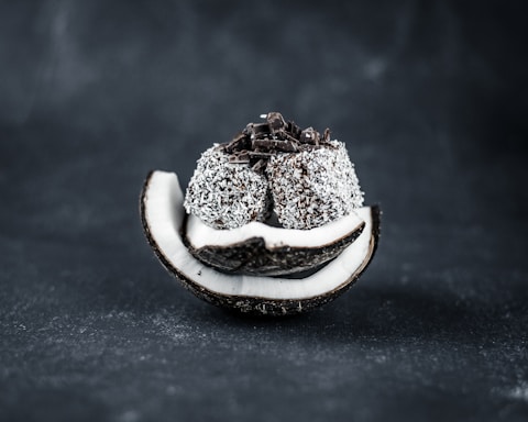 A sliced coconut shell filled with a stack of coconut-covered chocolate balls with chocolate shavings on top, set against a dark background.
