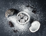 Coconut halves and chocolate-covered cubes are scattered on a dark surface with a generous dusting of desiccated coconut. The chocolate treats are nestled in a coconut shell, enhancing the theme of coconut and chocolate fusion.