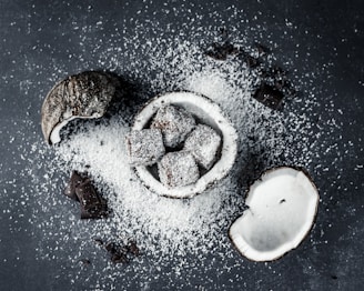 Coconut halves and chocolate-covered cubes are scattered on a dark surface with a generous dusting of desiccated coconut. The chocolate treats are nestled in a coconut shell, enhancing the theme of coconut and chocolate fusion.