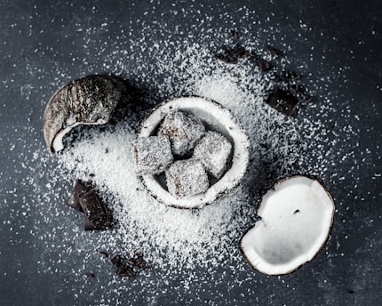 Coconut halves and chocolate-covered cubes are scattered on a dark surface with a generous dusting of desiccated coconut. The chocolate treats are nestled in a coconut shell, enhancing the theme of coconut and chocolate fusion.