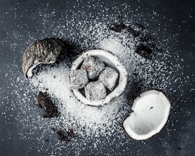 Coconut halves and chocolate-covered cubes are scattered on a dark surface with a generous dusting of desiccated coconut. The chocolate treats are nestled in a coconut shell, enhancing the theme of coconut and chocolate fusion.