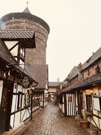 A cobblestone street lined with quaint, half-timbered houses and small shops leading to a large, round stone tower. The architecture has a medieval European style with ornate signs hanging from the buildings. Snow appears to be gently falling, adding a serene and nostalgic atmosphere.