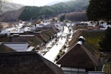 A quaint village with traditional thatched-roof houses is set in a mountainous area. A narrow path runs through the center, lined with people casually walking and exploring. Surrounding the village are forested hills with a mix of evergreen and deciduous trees.