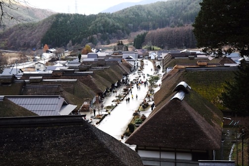 A quaint village with traditional thatched-roof houses is set in a mountainous area. A narrow path runs through the center, lined with people casually walking and exploring. Surrounding the village are forested hills with a mix of evergreen and deciduous trees.