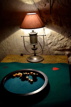 A rustic room setting features a vintage table lamp with a fabric shade, casting a warm glow on a stone wall. Below the lamp is a table with a circular tray filled with various small round objects, likely game pieces, lying on a green felt surface adorned with a red heart shape.