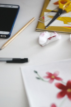 A cluttered desk features a smartphone, a pencil, a pen, a piece of tissue paper with a red stain, a pair of yellow scissors, a closed notebook with white and yellow paper on top, and a partially visible drawing of red and pink flowers.