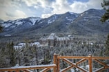 A large, historic hotel sits amidst a snowy forest with majestic mountains in the background. The wooden fence in the foreground adds a rustic touch to the scene. The sky is partly cloudy, and the setting conveys a sense of tranquility and grandeur.