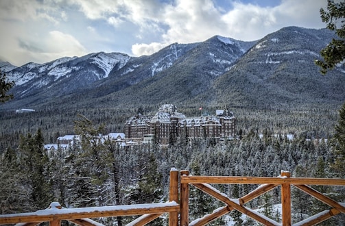 A large, historic hotel sits amidst a snowy forest with majestic mountains in the background. The wooden fence in the foreground adds a rustic touch to the scene. The sky is partly cloudy, and the setting conveys a sense of tranquility and grandeur.