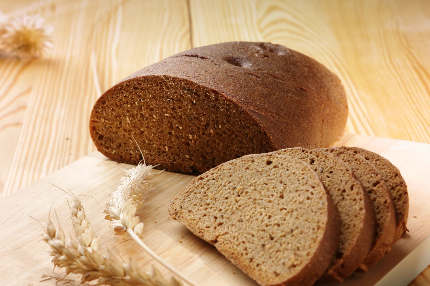 A dark rye loaf cut into slices on a board, loose wheat ears beside it